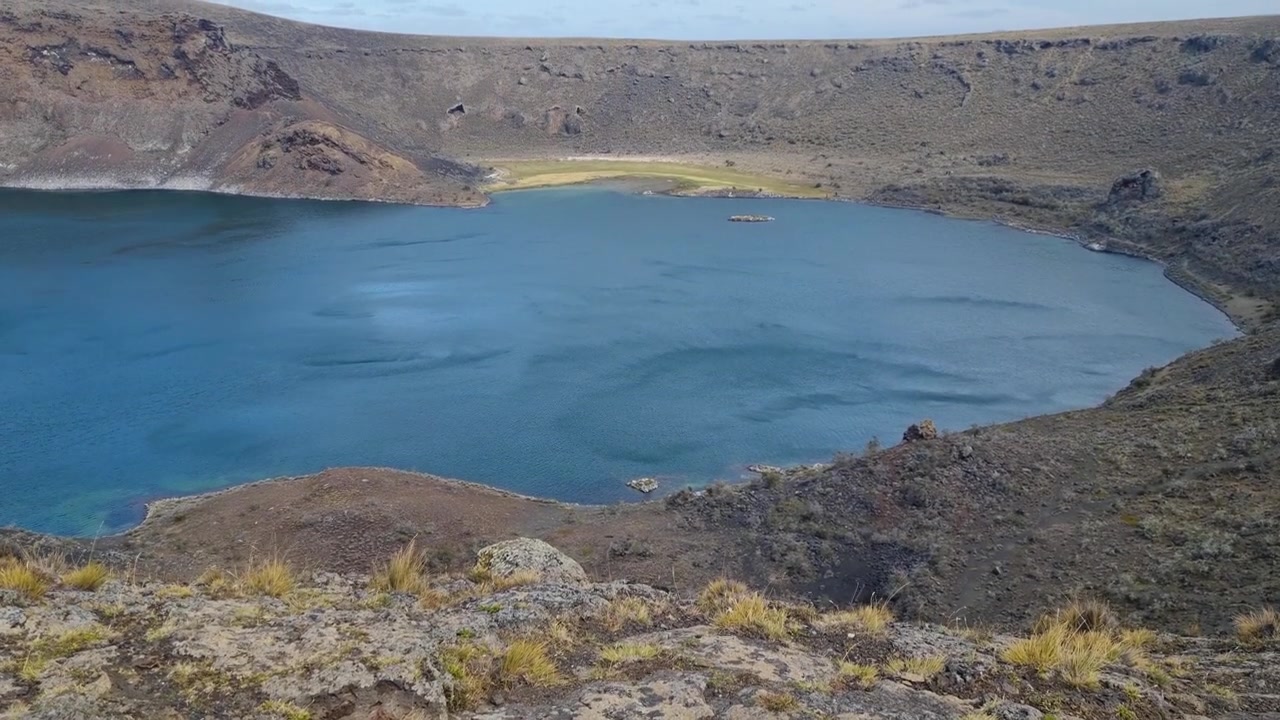 Aguas de un azul profundo de Laguna Azul se ondulan bajo fuertes vientos patagónicos en el campo volcánico de Pali Aike, cerca de Río Gallegos, Argentina, mostrando un espectacular lago de cráter volcánico y una belleza remota