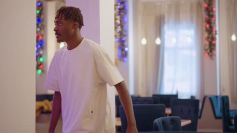 a man with braided hair in white shirt arranges set of blue chairs in spacious, decorated room with warm light and festive atmosphere, preparing for event or gathering