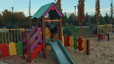 boy wearing jacket and cap waving on the horizontal bar. kid sits down on the slide and taps his feet happily. playground backdrop.