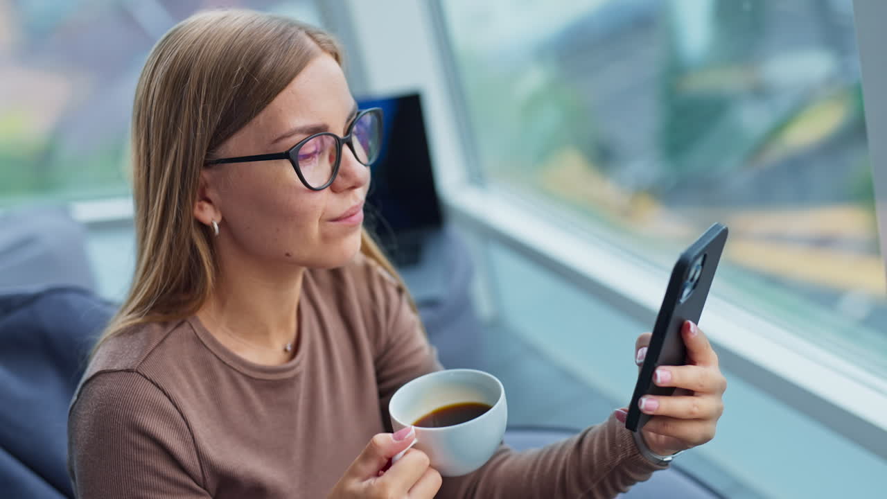 Smiling young blonde woman drinking coffee looking at her phone. Lady having lunch break sitting in bean bag chair near window. High angle view.