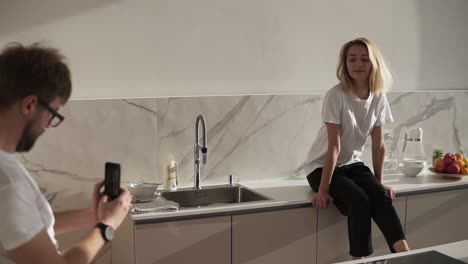 Happy-couple-cooking-at-modern-kitchen-together.-Smiling-woman-is-sitting-on-a-counter-while-her-husband-making-photo-on-mobile-phone.-Joyful-wife-posing-for-her-man.-Wearing-domestic-clothes