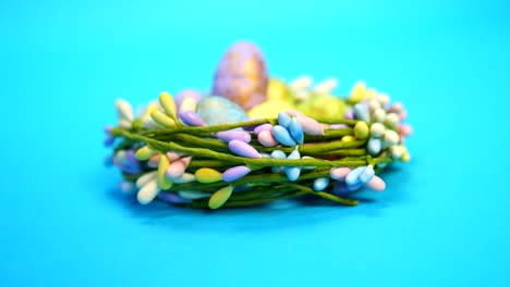 Handaufnahme-Von-Bunten-Ostereiern,-Die-In-Einem-Blumennest-Vor-Blauem-Hintergrund-Gehalten-Werden