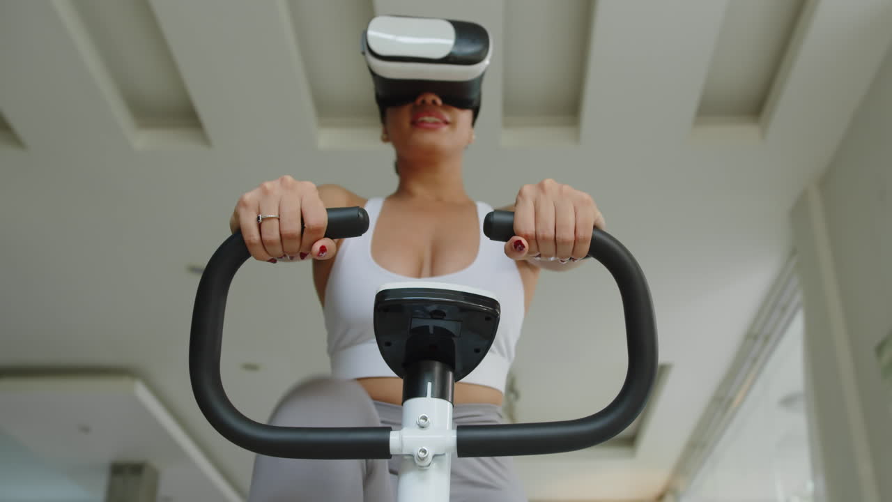 Female Athlete Watching Video in VR Headset during Exercising on Fitness Bike