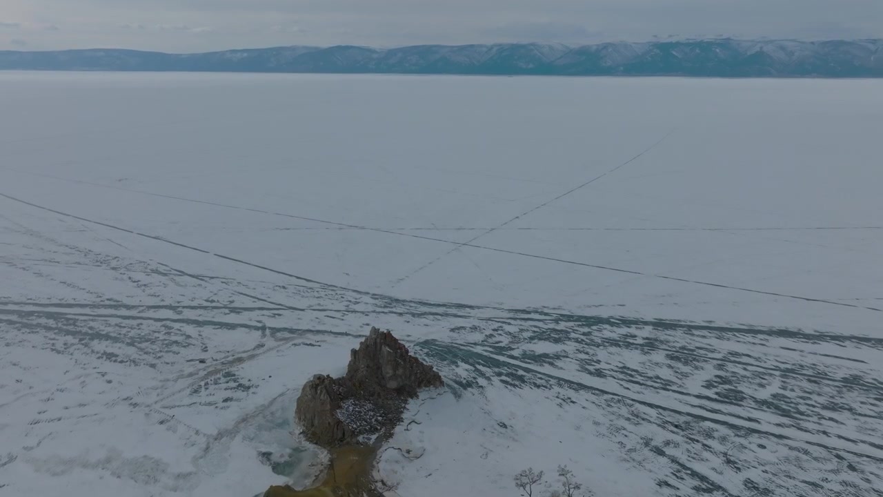 Droneoptagelser af en klippefyldt holm omgivet af revnet is på den frosne Bajkalsø i Irkutsk oblast, Rusland, med fjerne bjerge under en overskyet himmel
