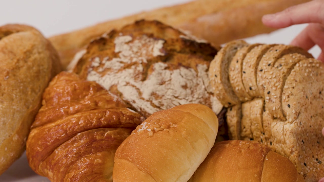 Close-up of hands placing sliced multigrain bread among croissants and baguettes on white background.