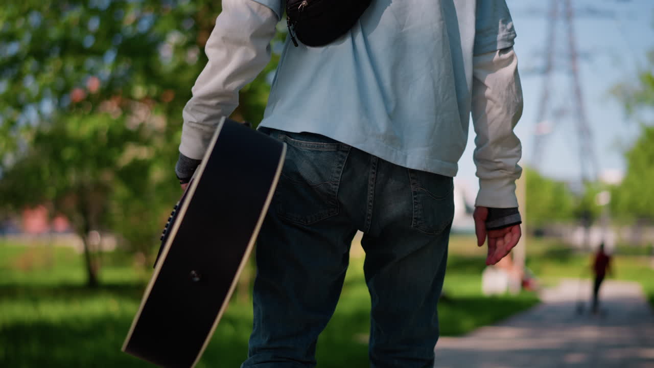 young male guitarist walking in park carrying guitar case, strolling along sunlit pathway with backpack, casual jeans and hoodie, acoustic case swinging by side, peaceful summer afternoon, musician