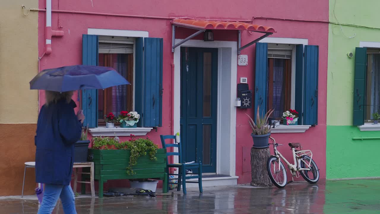Woman with umbrella on rainy day walks past painted pink house Burano