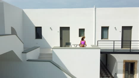 a woman wearing a straw hat and a pink bathing suit sits on the balcony of the white mansion