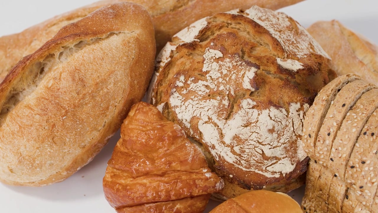 Slow camera zoom on variety of crusty sourdough, baguettes, and croissants against white backdrop.