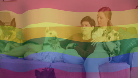 animation of pride rainbow flag over happy caucasian lesbian couple relaxing on couch with pet dogs