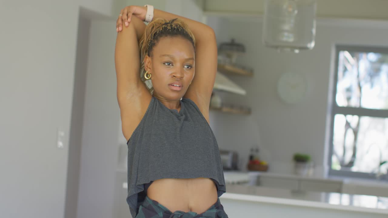 Donna afroamericana in crop top che si allunga in una cucina illuminata dal sole, alzando, incrociando e afferrando il gomito.