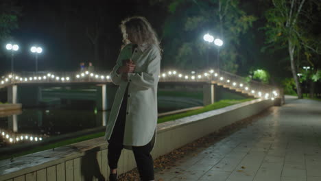 woman in white coat walks to wooden fence by river under glowing bridge lights at night, preparing to sit, cross legs, and read book in quiet, scenic outdoor environment