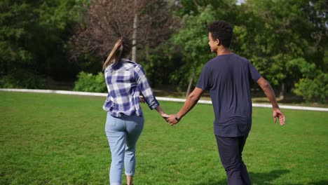 Back-view-follow-shot-of-cute-attractive-international-couple-holding-hands-and-running-on-the-field-with-green-grass-in-park