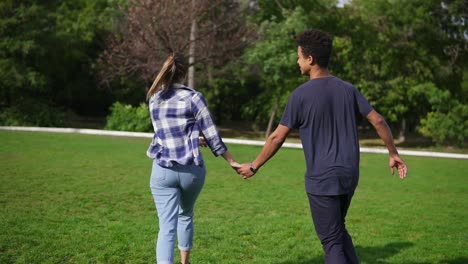 vista trasera seguir la toma de una pareja internacional linda y atractiva cogida de la mano y corriendo en el campo con hierba verde en el parque