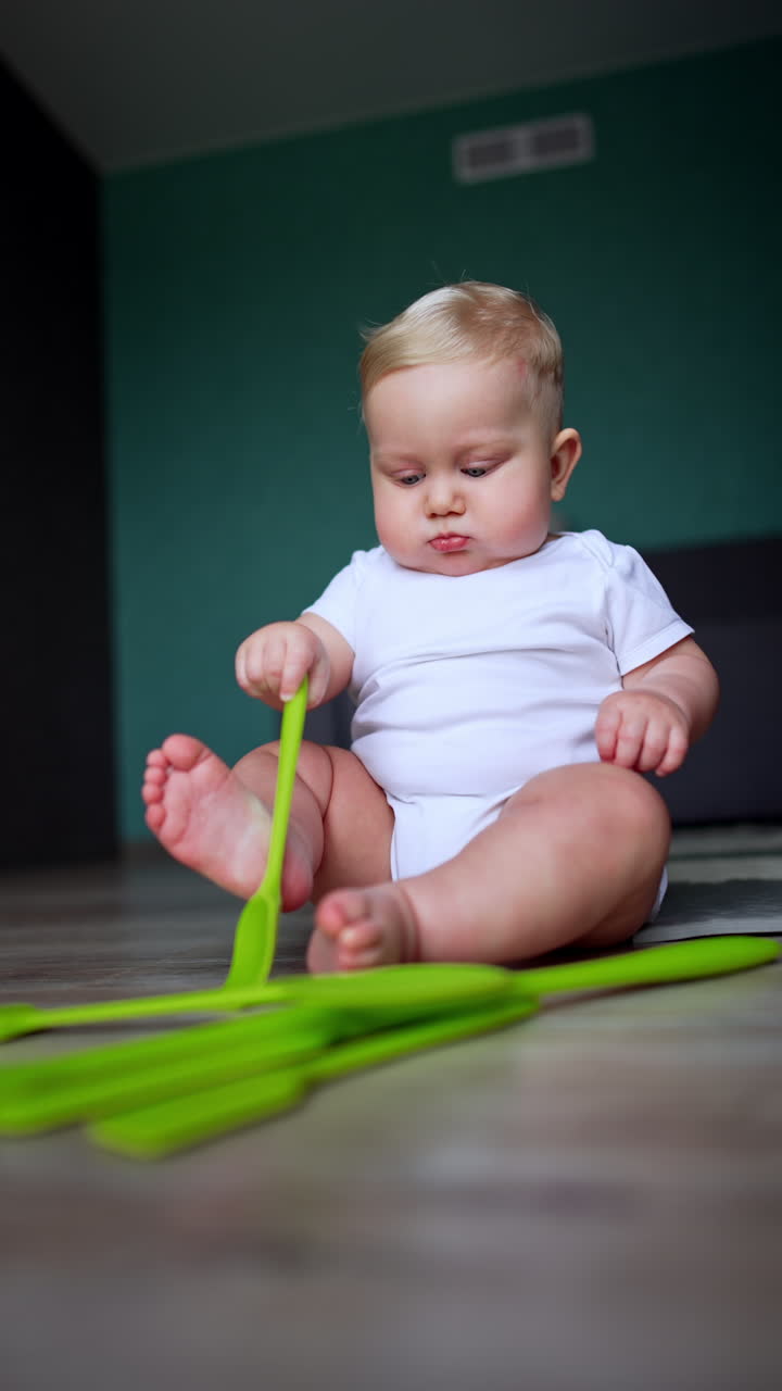 Lovely plump baby in white bodysuit sits on the floor. Cute child plays with kitchen utensils. Low angle view. Vertical video.