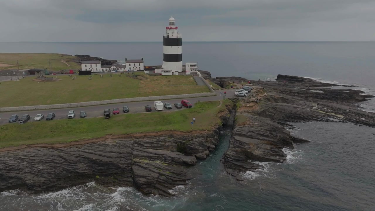 Et luftfoto af det historiske sort- og hvidstribede Hook Fyr, beliggende på en barsk klippefyldt halvø i County Wexford, Irland, under en overskyet himmel.