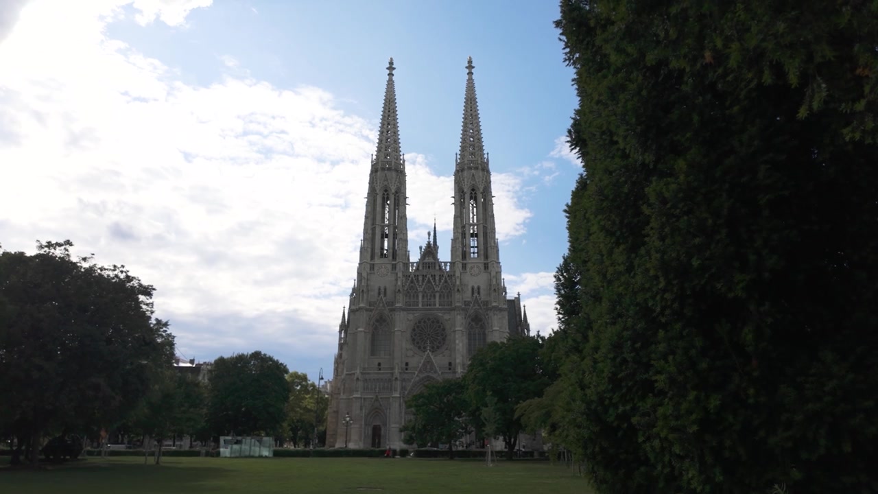 Votivkirken i Wien, set fra en nærliggende park, omgivet af træer og en overskyet himmel.