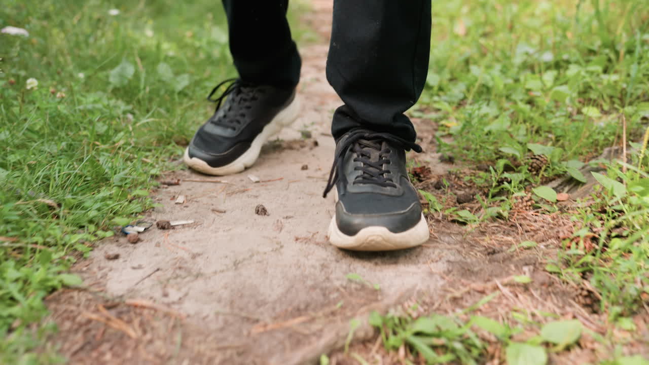 Leg view of man in black trousers and canvas sneakers walking slowly on narrow forest pathway surrounded by greenery, grass, and soil under calm natural daylight outdoor atmosphere