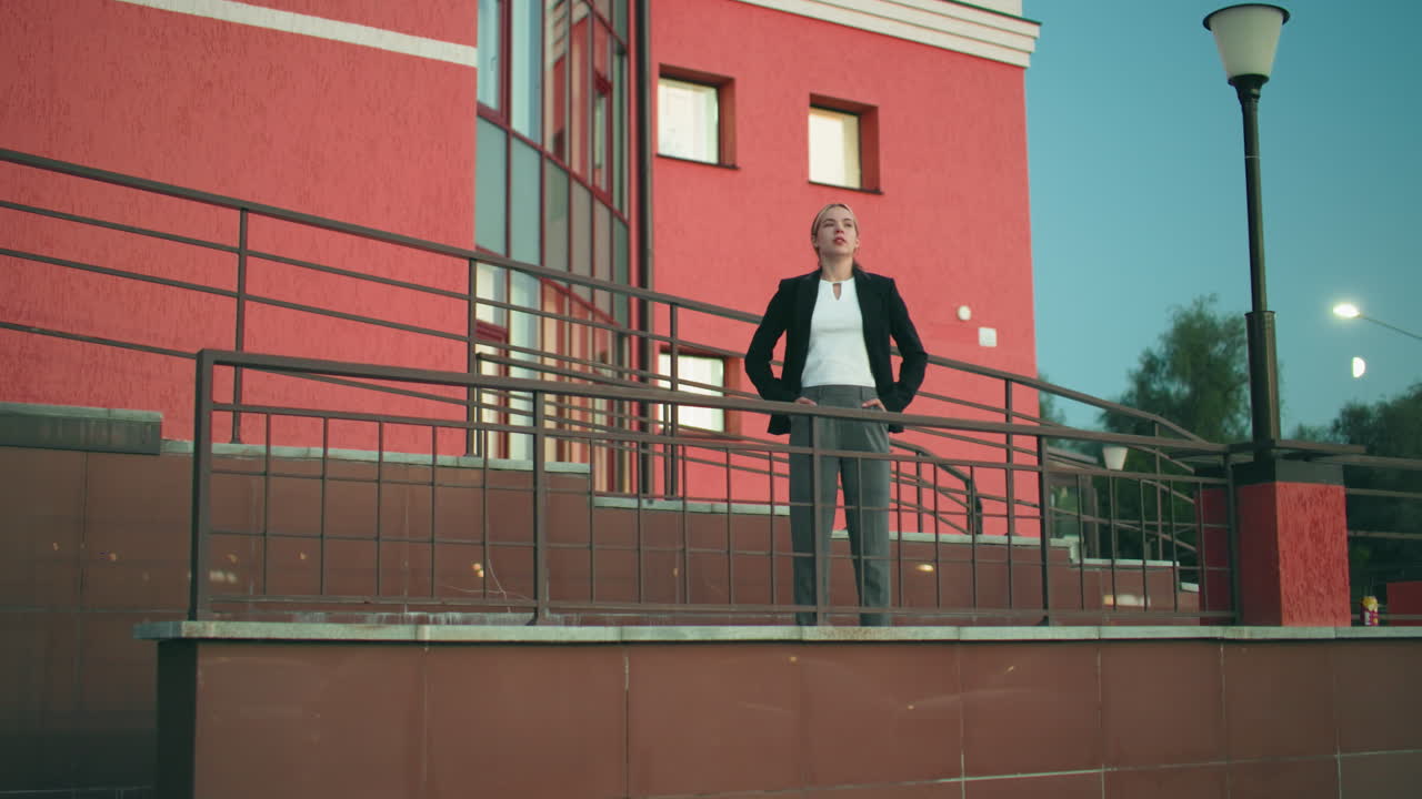 Confident woman in black blazer and white top poses with hands in pockets in front of residence, iron rail and street light nearby, reflection visible on tiled wall, tree in background