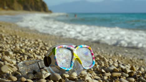 gafas de snorkel para niños en la playa de guijarros con olas de mar salpicando en el fondo, concepto de vacaciones