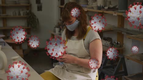 Animation-of-covid-19-cells-over-woman-wearing-face-mask-using-smartphone-in-pottery-workshop