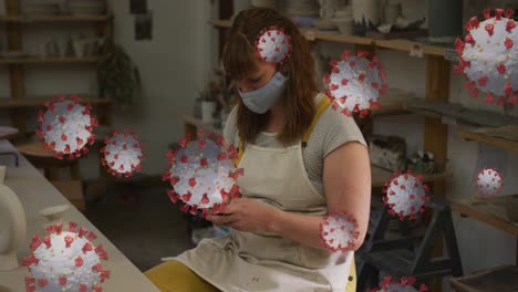 animation of covid 19 cells over woman wearing face mask using smartphone in pottery workshop