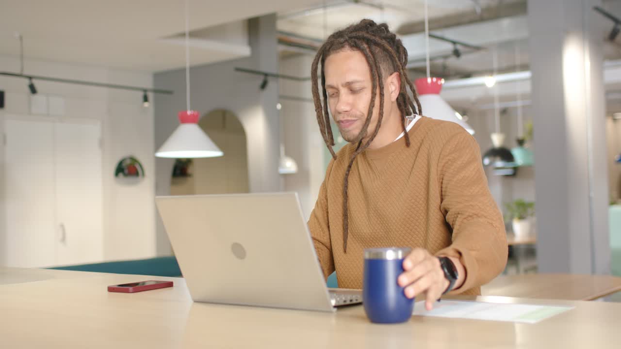 Dreadlocked man in brown sweater at office sipping blue mug to focus, then typing on laptop