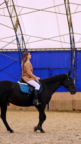 horse riding inside the drill-hall. long-haired woman in beige jacket and white pants rides a beautiful calm black horse. vertical video