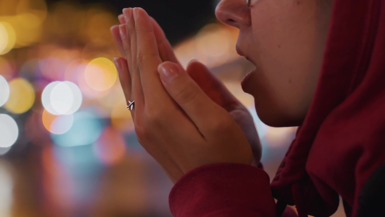 Primo piano notturno sul lungomare: mani che si scaldano vicino a luci bokeh. Un pendolare romantico si ferma accanto al fiume, anello visibile, un leggero battito di mani. Luci sfocate del ponte si riflettono sull'acqua, una comoda felpa rossa con cappuccio, una serata intima