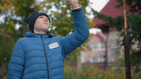 boy in blue jacket and beanie holds a leaf high, inspecting it against vibrant autumn foliage and rustic background, thoughtful expression and bright greenery evoke a sense of nature exploration