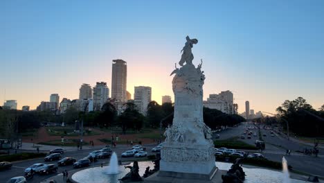 aerial slow pull out shot capturing monumental sculpture carta magna and four regions of argentina at rotary road with peak hour traffics moving and downtown cityscape in the background