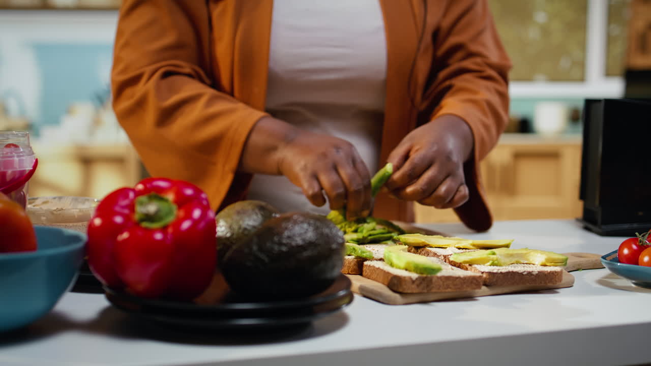 Vlogger di cucina afroamericano che spalma fette di avocado su pane tostato per una scelta salutare.