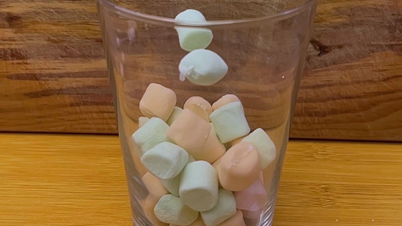 Close-up de marshmallows coloridos caindo em um copo transparente sobre uma mesa de madeira, capturando pedaços de doce macio em movimento com uma composição interna simples