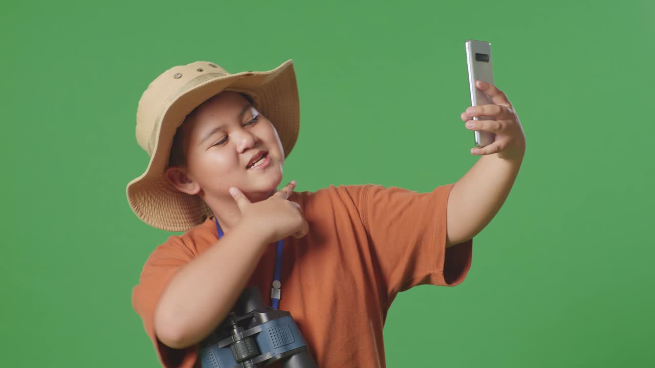 모자와 안경을 쓴 아시아 관광 소년이 초록색 스크린 배경에 서서 스마트폰으로 셀피를 찍습니다. 소년 연구자는 무언가를 조사합니다.