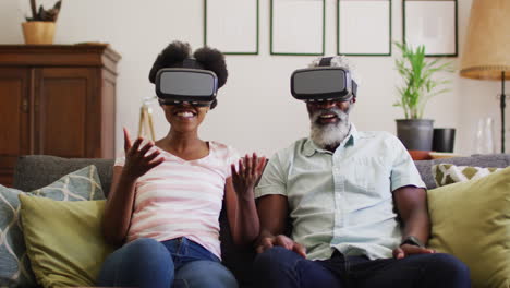 happy african american couple sitting on couch wearing vr headsets pointing and smiling