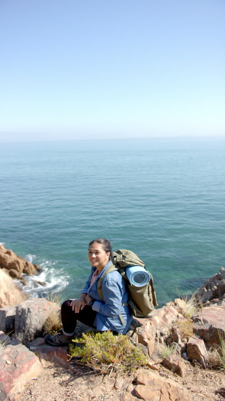 Vertical video: Hiking with backpack and yoga mat, woman sitting on cliff by ocean
