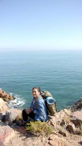 vertical video: hiking with backpack and yoga mat, woman sitting on cliff by ocean