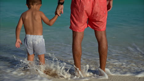 Cámara-Lenta-De-Un-Pequeño-Niño-Latino-Mexicano-Sosteniendo-La-Mano-De-Su-Padre-Parado-En-La-Playa-Esperando-Que-Las-Olas-Golpeen-Sus-Piernas