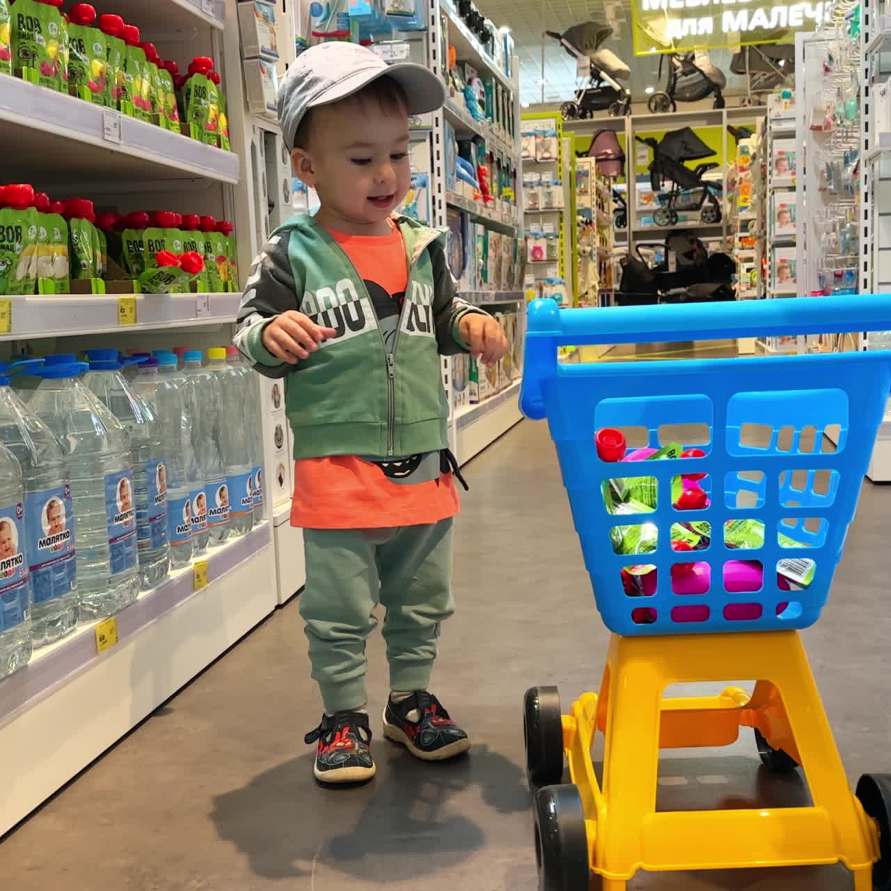 Cheerful baby boy in a cap throws product from a shelf to his cart. Little child shopping in a grocery store