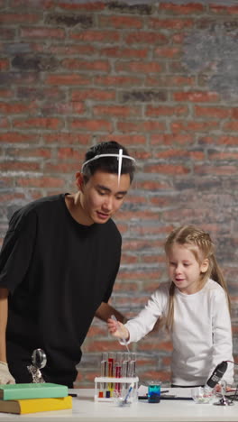 pretty blonde little girl drops colored liquid into beaker doing interesting test with young asian teacher help at chemistry lesson in studio