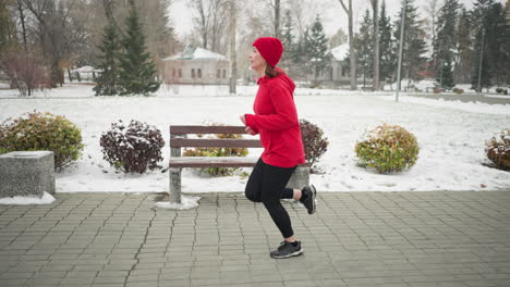 side view of tired woman jogging along snowy park pathway in red hoodie and black leggings, surrounded by benches, vibrant bushes, frosty winter trees, and distant snow-dusted houses