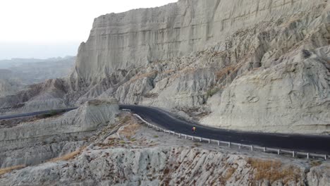 Aerial-View-Of-Cyclist-Riding-On-Remote-Road-Beside-Dramatic-Rock-Cliff-On-The-Makran-Coastal-Highway