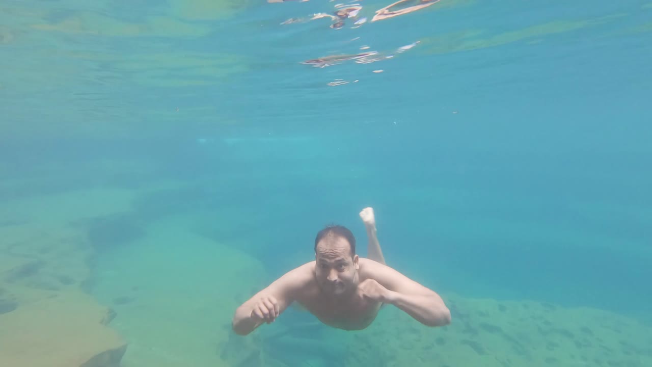 young man swimming in blue crystal clear under water of waterfall from top angle