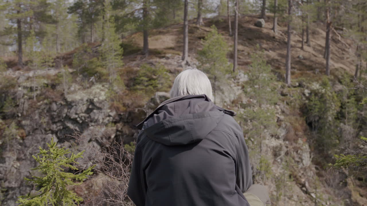 Older man sitting quietly on rocky edge facing rugged forest hillside.