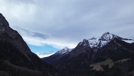 drone slowly moving above pine forest with snow covered summits in background in vorarlberg, austria
