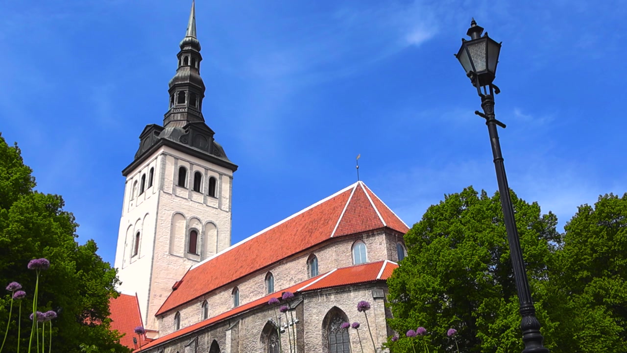 Il museo Niguliste, un'ex chiesa medievale a Tallinn, Estonia, in una soleggiata giornata estiva, con alberi verdi, fiori di allium viola e un lampione d'epoca contro un cielo azzurro limpido.