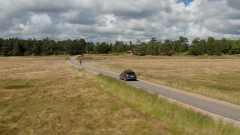 Fly-around-car-driving-on-narrow-road-in-countryside.-Cyclist-riding-in-front-of-car.-Buildings-on-edge-of-forest-under-dramatic-clouds-in-sky.-Denmark