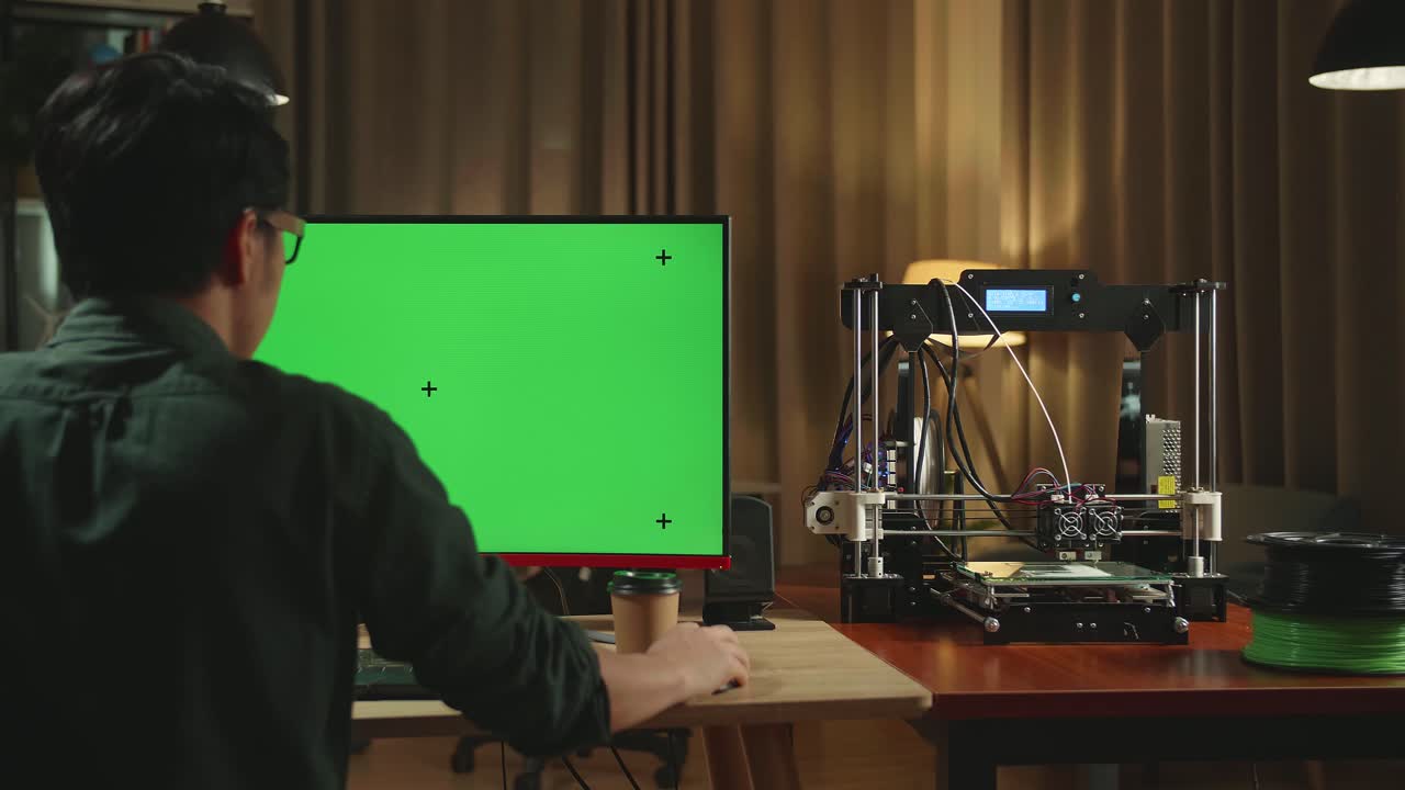 Back View Of Asian Man Work On 3D Printer And Personal Computer With Mock Up Green Screen Display In Home Office