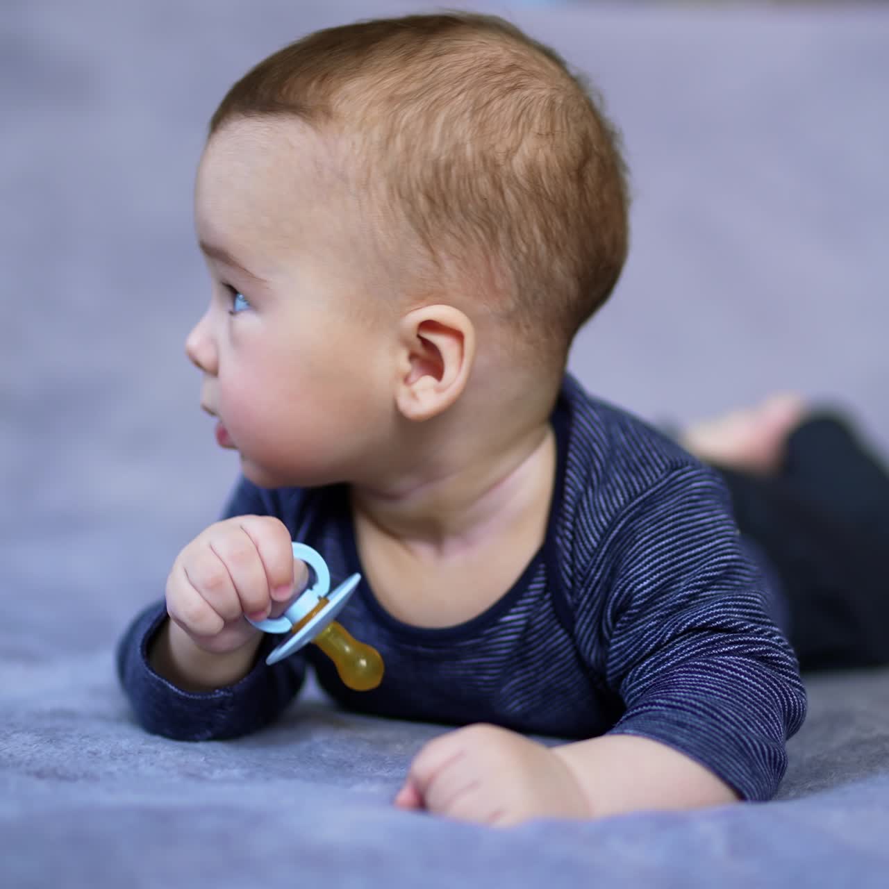 Lovely little kid lies on bed and looks up surprised. Cute toddler boy tries to shove the pacifier into a mouth. Grey blurred backdrop