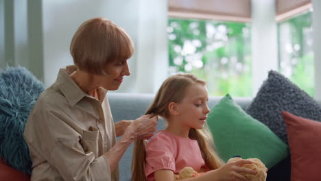 Fröhliche-Großmutter,-Die-Ihrer-Kleinen-Enkelin-Im-Wohnzimmer-Die-Haare-Flechtet.